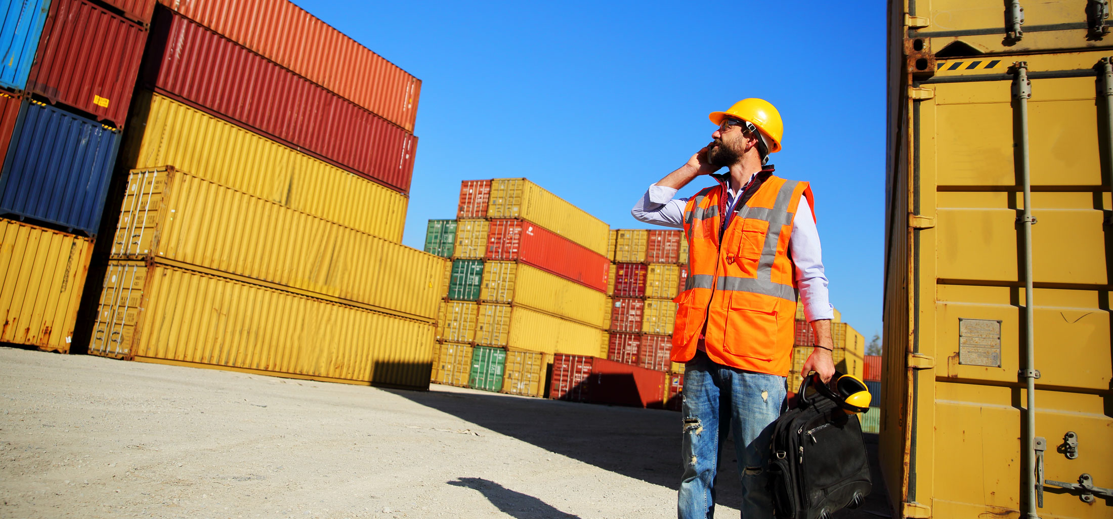 Customs officer on the phone while standing amongst shipping freight containers at a dock yard.
