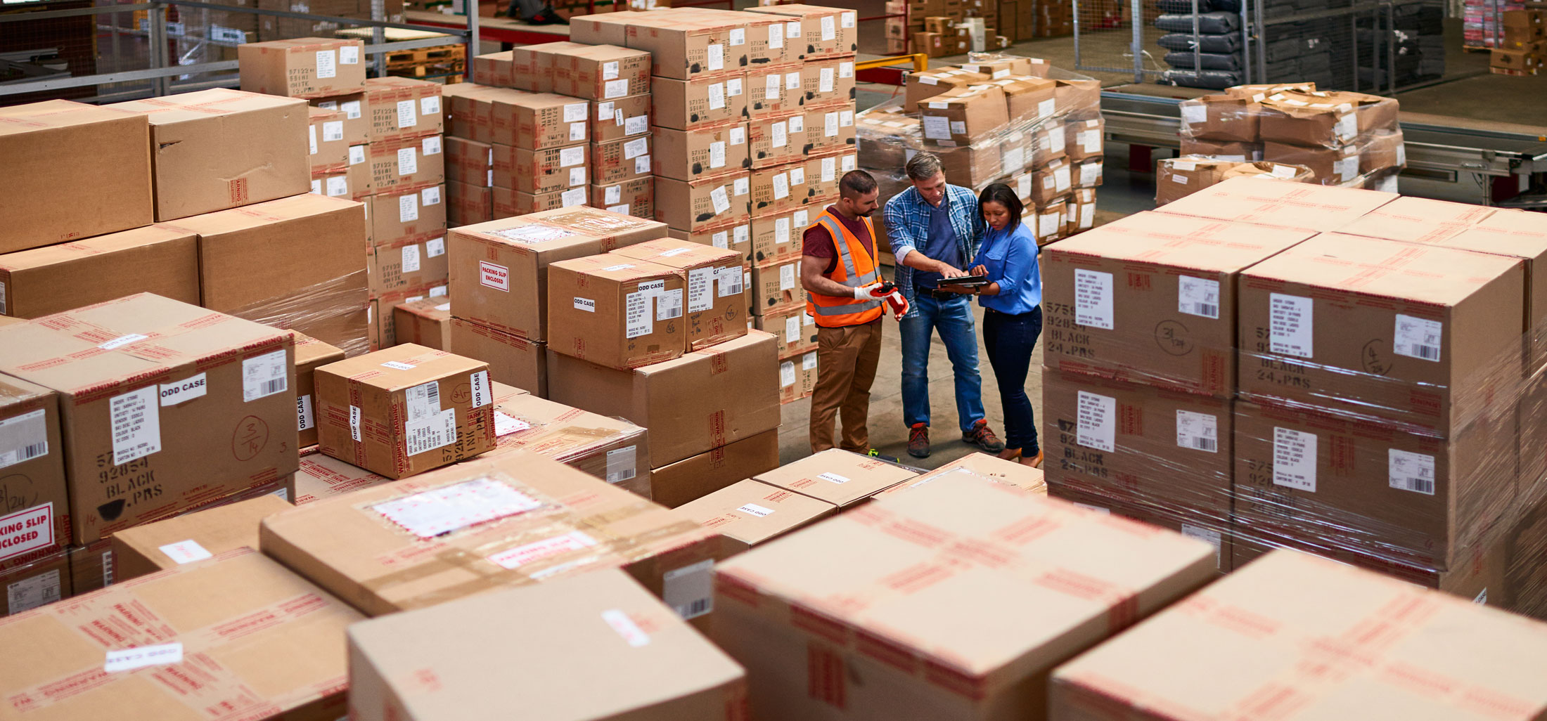 Interior of warehouse with stacked boxes and 3 logistics personnel looking at tablet device